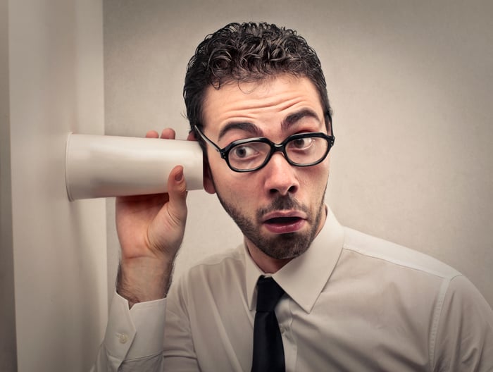 A man in a shirt and tie listens through a wall using a plastic cup.