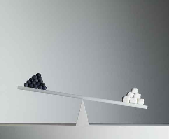 A seesaw with a pyramid of small black spheres on the left and larger white cubes on the right, against a blank gray wall.