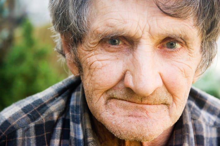 An elderly man in a flannel shirt pensively smirking. 