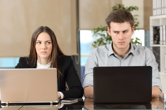 Male and female at laptops sitting side by side and glaring at one another