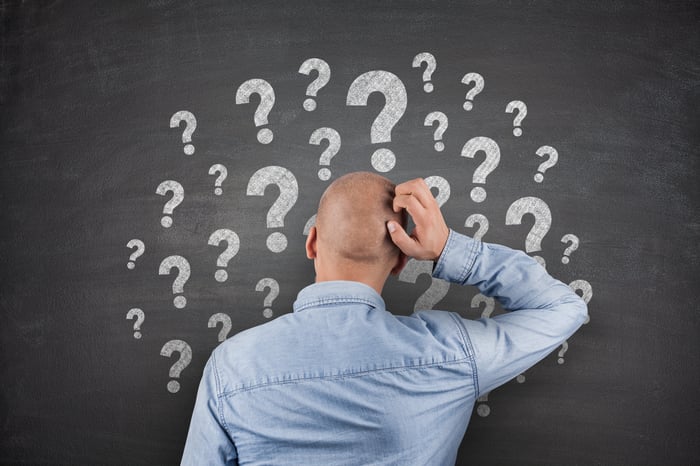 Man scratching his head in front of question marks on a blackboard. 