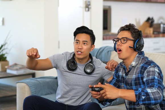 Two young men sitting on a sofa -- one playing a console video game and wearing Turtle Beach headsets and the other cheering him on.