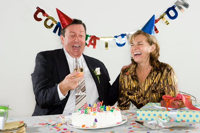 Two middle aged people sitting behind a retirement party cake, wearing party hats and smiles