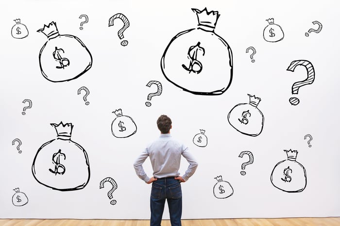 Man with hands on hips looking at wall covered with drawings of money bags and question marks.