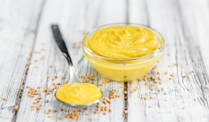 A small bowl of mustard and a spoon on a wooden table, strewn with mustard seeds