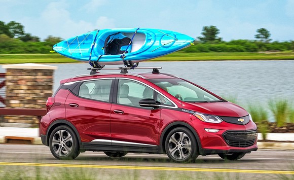 A red Chevrolet Bolt EV, an upright small hatchback, with a kayak on its roof, driving near a lake.