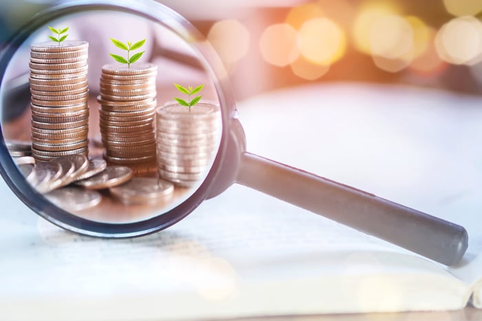 View through magnifying glass showing stacks of coins topped by sprouting plants.