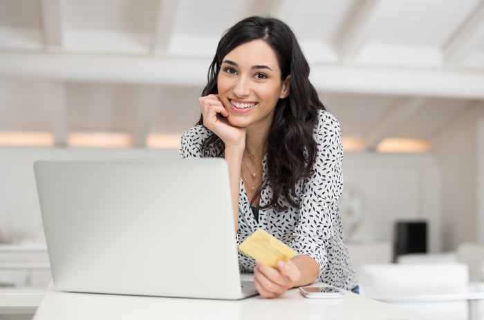 A smiling young woman holding a credit card in her left hand while in front of her laptop.