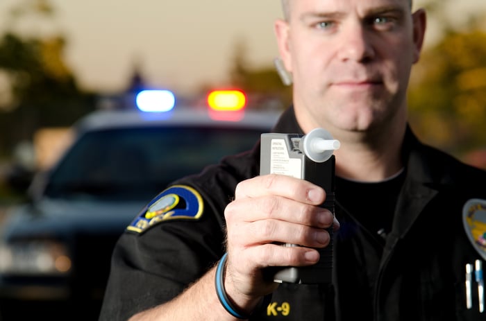 A police officer holding a breathalyzer device, with his patrol vehicle in the background.