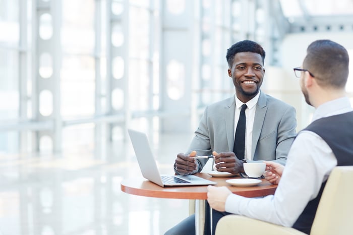 Two professional men talking at a small table with a laptop, with one man smiing