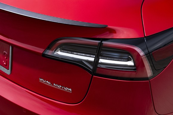 A close-up of the "Dual Motor" badge on the back of a red Tesla Model 3 sedan