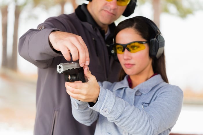 Woman holding pistol and wearing safety glasses and earmuffs. Another person holds the top of the firearm