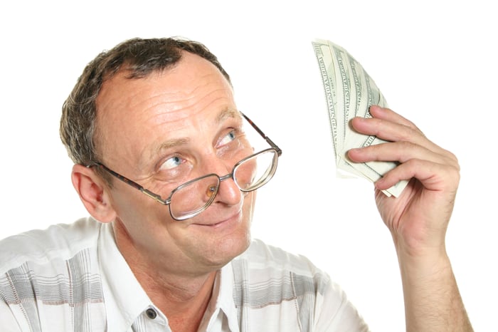 A baby boomer with glasses looking up at a small stack of cash bills he's holding in his left hand.