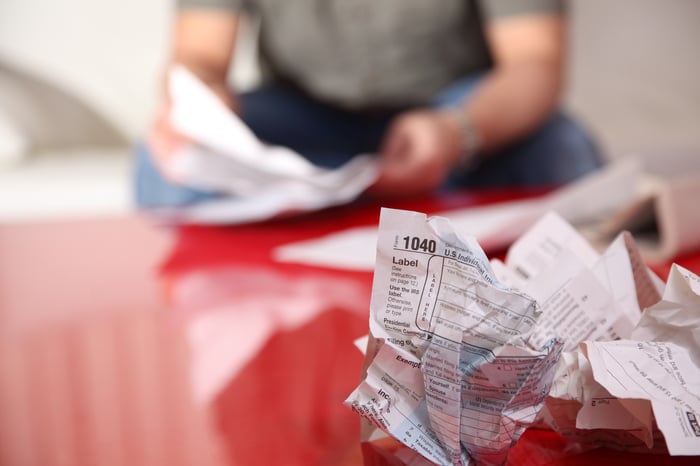 A crumpled up tax form in the foreground on a table, with a man looking over his tax documents in the background.