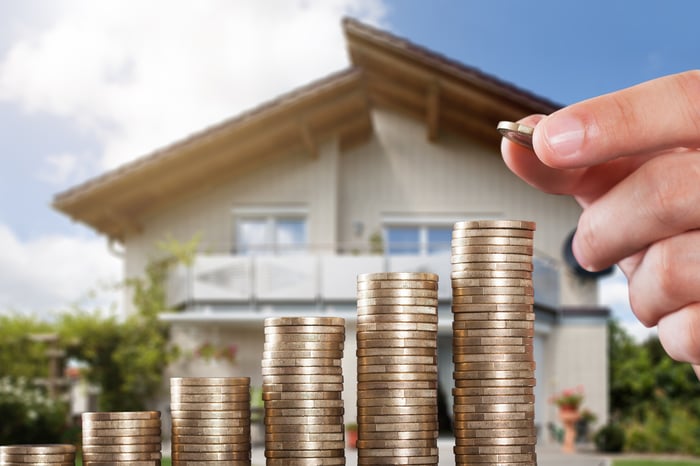 An ascending stack of coins in front of a house with a hand adding another coin to the highest stack. 