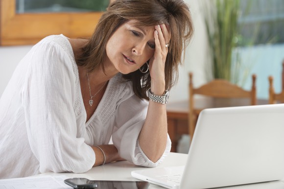 A worried-looking woman sitting in front of an open laptop with her left hand on her forehead.