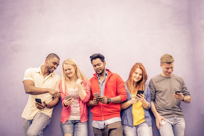 FIve adults leaning on a wall with smartphones in their hands