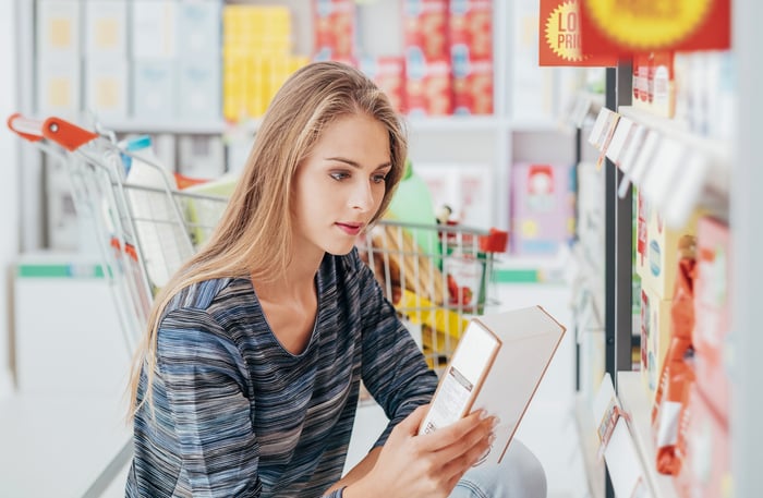 Woman crouching beside shopping cart in a discount store to read a product label.