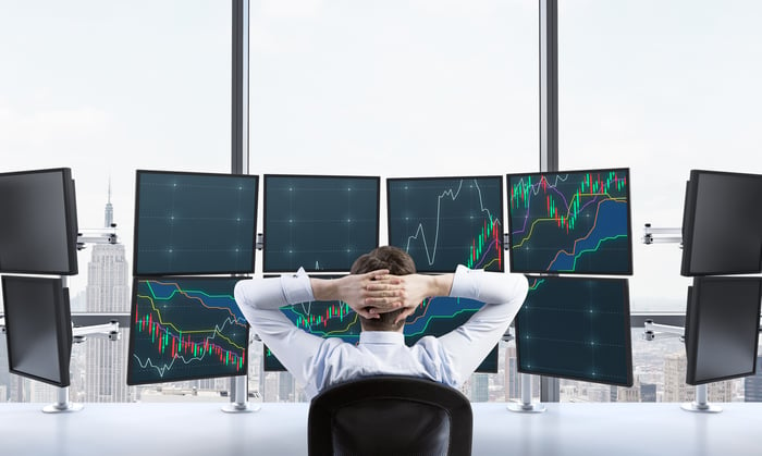 A man sitting with his fingers interlaced behind his head watching a bank of monitors showing graph lines.