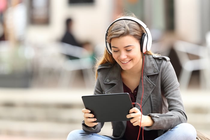 A girl sitting wearing headphones smiling and looking at her tablet.