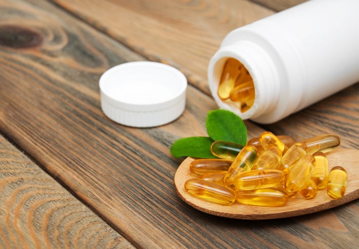 A bottle of softgel capsules tipped over on a table, with about a dozen capsules piled in front of the bottle.