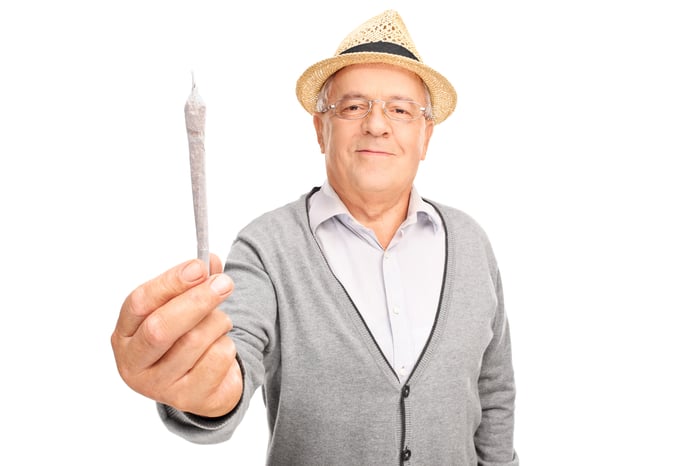 A baby boomer holding a rolled cannabis joint with his arm outstretched.