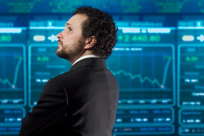 An investor in a suit, looking at various digital charts on a large ticker board.