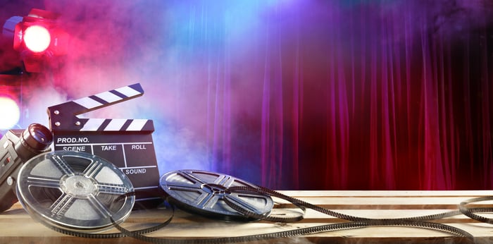 A clapperboard and film reels in front of smoke effects, a spotlight, and a theater curtain