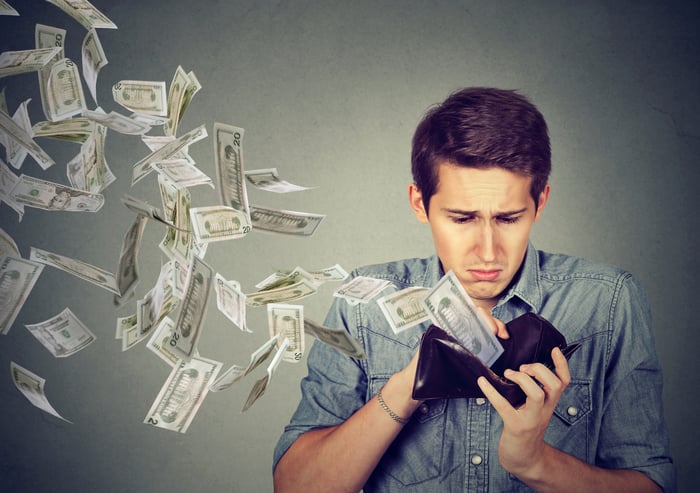 A young man looks sadly at his open wallet as paper American currency flies out.