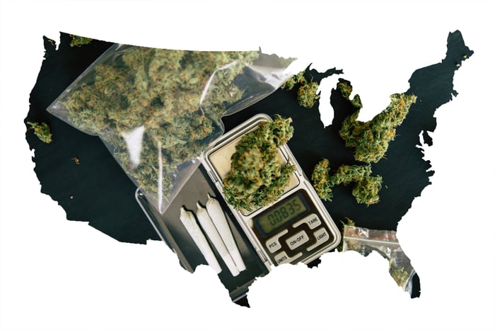 A dark outline of the U.S., mostly filled in with dried cannabis baggies, rolled joints, and a scale. 