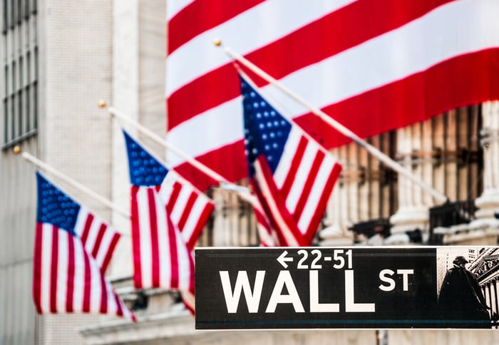 The facade of the NYSE draped in a large American flag, with the Wall Street street sign in focus.