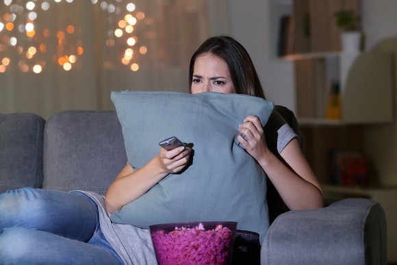 A young woman, hugging a pillow tight with the TV remote in hand a a popcorn bucket close at hand.
