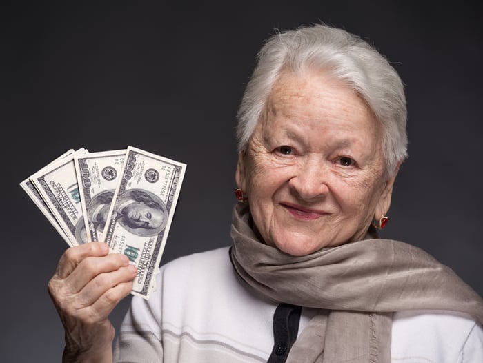 A smiling older woman is holding some hundred dollar bills, fanned out.