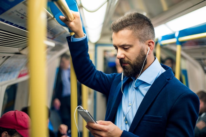 Man with headphones looking at his phone and holding onto train pole.