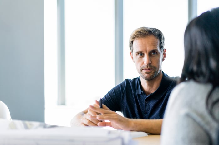 Man with a serious expression and a collared shirt sitting across from a woman in an office