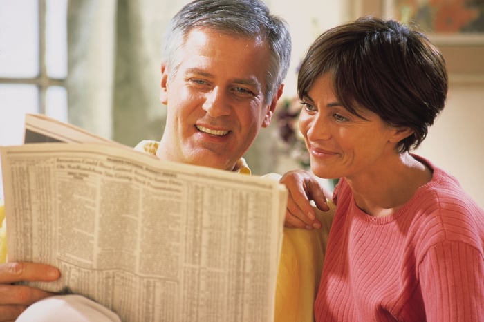 A smiling married couple reading the financial portion of a newspaper. 