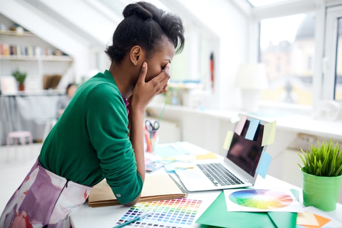 A young woman sitting at desk in front of laptop with her face in her hands.