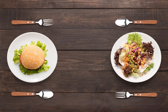 A burger on a plate flanked by a fork and spoon facing a salad on a plate flanked by a fork and spoon