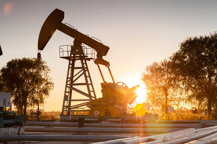 An oil pump with pipes in the foreground and the sun setting behind it.