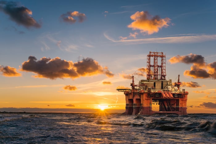 A drilling rig in the water with the sun setting in the background.