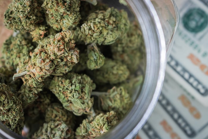 A jar filled with cannabis buds that's lying atop a fanned pile of twenty dollar bills.