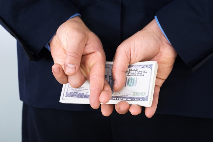 A businessman in a suit holding a neat stack of cash behind his back while also crossing his fingers.