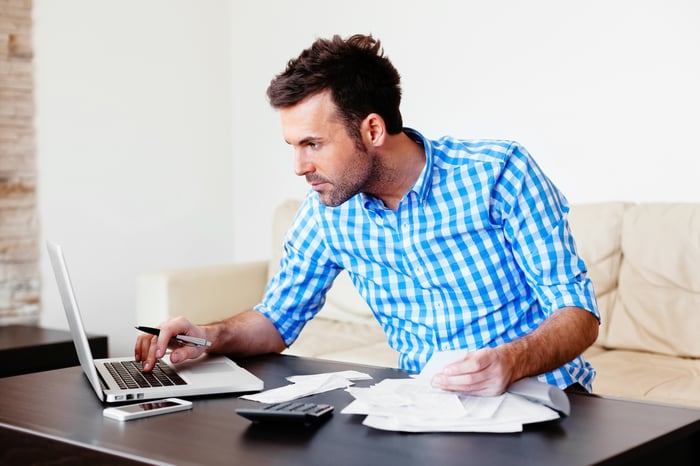 A man sits at a desk with a calculator, a pen in hand, and a pile of papers, while looking at a laptop screen.