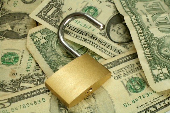 An open padlock sits on top of a scattered pile of U.S. currency.