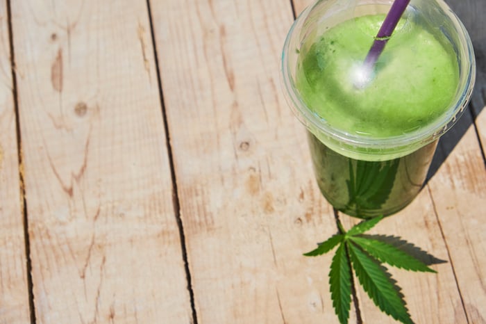 Glass filled with a green beverage sitting on a wood surface with a canabis leaf next to it. 