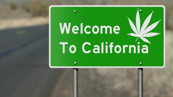 A green highway sign with white lettering that reads, Welcome to California, with a white cannabis leaf in the top right corner. 