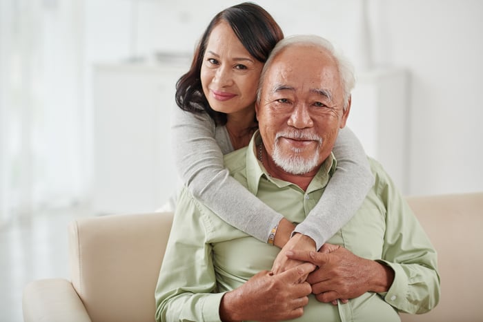 A happy senior couple -- the man is sitting on a couch and the woman is embracing him while he holds her hands.