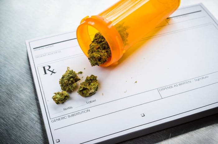 A tipped over prescription bottle containing a few dried cannabis buds that's lying atop a doctors prescription pad.
