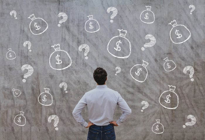 A man looking at a chalkboard covered by drawings of money bags and question marks.