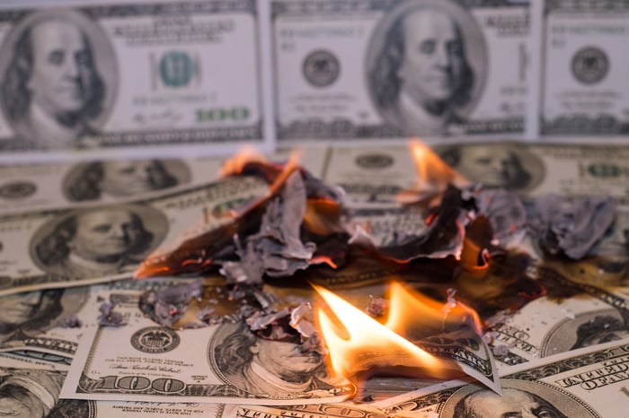 A small pile of hundred dollar bills burning, with multiple hundred dollar bills laid out as wallpaper in the background.